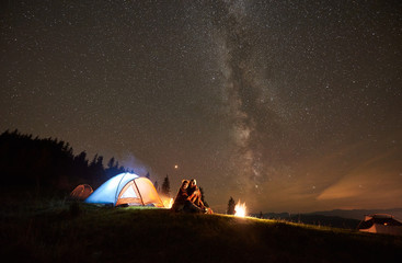 Night summer camping in the mountains. Happy couple hikers sitting together beside bonfire and glowing tourist tent. On background forest and beautiful night starry sky full of stars and Milky way.