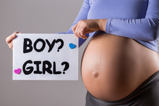 Image of close up stomach of pregnant woman holding paper with questions  boy or a girl on gray background.