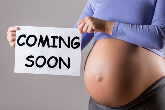 Image of close up stomach of pregnant woman holding paper with text coming soon on gray background.