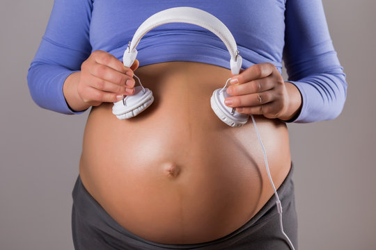 Image of close up stomach of pregnant woman holding headphones on gray background.