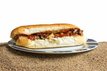 rolled sausage (South African Borewores roll) on a roll woth tomatoe relish and onions on a plate on beach sand, isolated on a white background