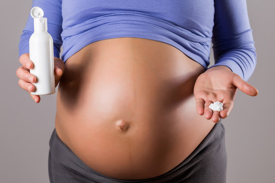 Image of pregnant woman applying moisturizer on her stomach on gray background.