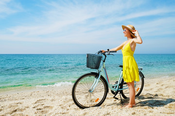 Fototapeta premium Portrait of young carefree woman in bright yellow dress with bicycle at ocean beach. Female wearing broad brim straw hat biking on sandy sea shore on sunny day. Copy space, background.