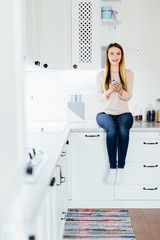 Young blonde woman using mobile phone sitting in modern kitchen