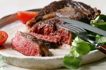 Grilled roasted sliced medium rare beef steak served in ceramic plate with green field salad, cherry tomatoes, cutlery, over white marble table. Close up