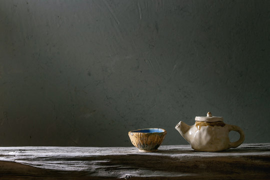 Craft handmade ceramic teapot and cup for tea ceremony standing on old wooden shelf in dark room.