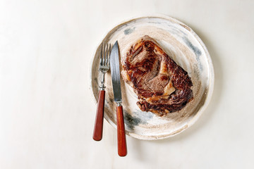 Grilled roasted medium rare beef steak served in ceramic plate with cutlery over white marble background. Flat lay, space
