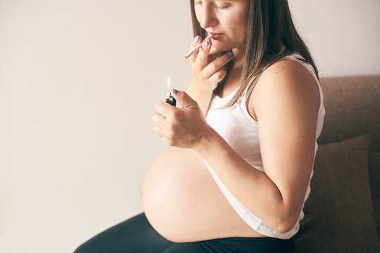 Close up view of lighter and cigarette in hands of pregnant woman. Young future mother in white shirt smoking during pregnancy period. Concept of threat and harmfulness.