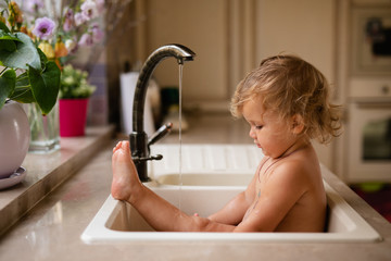 Baby taking bath in kitchen sink. Child playing with foam and soap bubbles in sunny bathroom with...