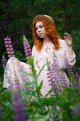 A beautiful red-haired woman stands and poses in a field of purple lupins and enjoys the view