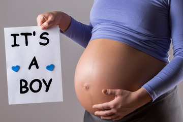 Image of close up stomach of pregnant woman holding paper with text it's a boy on gray background.