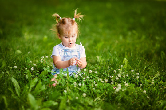 Portrait of adorable little blonde girl lying on grass and propping up her face at summer green park. Baby girl with white flowers