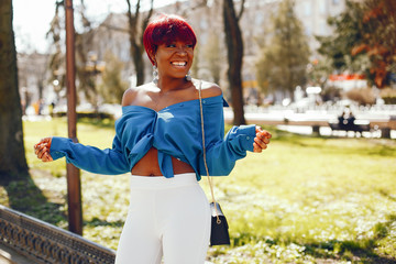 beautiful and stylish dark-skinned girl with red short hair, dressed in a blue top with long sleeves and white pants standing in a summer sunny park