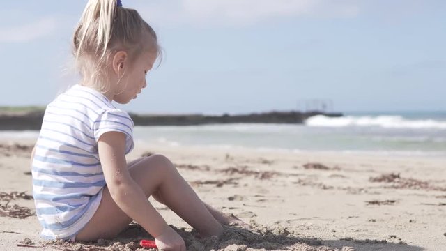 Beautiful girl playing on the sandy beach by the sea. The sea is not calm, a lot of waves. The child buries its feet with sand. She diligently plays her game