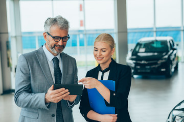 salesman showing woman online car gadgets in car dealership showroom