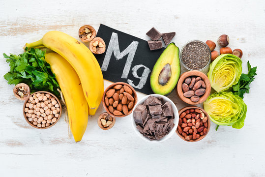 Foods containing natural magnesium. Mg: Chocolate, banana, cocoa, nuts, avocados, broccoli, almonds. Top view. On a white wooden background.