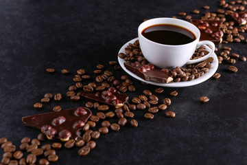 Scattered coffee grains, a cup and black chocolate on a black stone table. Copy space.