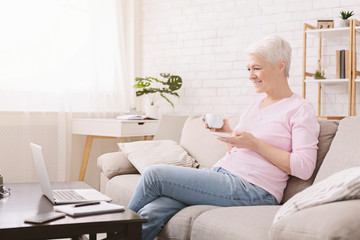 Positive senior woman enjoying coffee and watching news on laptop