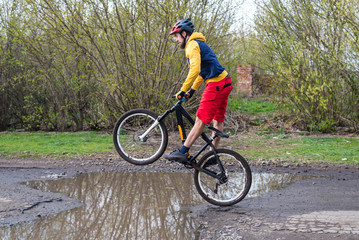 Fototapeta na wymiar A cyclist in red shorts and a yellow jacket riding a bicycle on the rear wheel through a puddle.