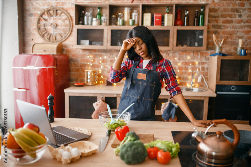 Wall mural sleepy black woman in apron cooking on the kitchen