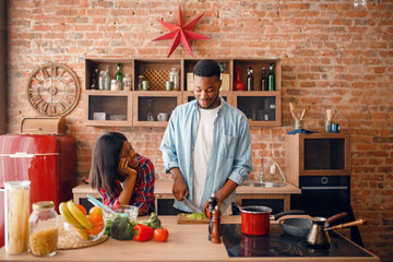 Black man cooking on kitchen, wife drinks coffee