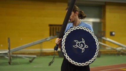 Young man with long hair demonstrates LARP foam combat holding a shield and a sword inside a gym. 4k.