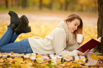 A beautiful girl reads a book and drinks warm tea, hot shikolad, coffee in a warm autumn park. Concept of autumn warmth, atmosphere and comfort. With space for an inscription