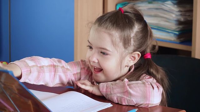 Cheerful little girl smiling and reading book sitting at desk at early development school in close up. Child in kindergarten