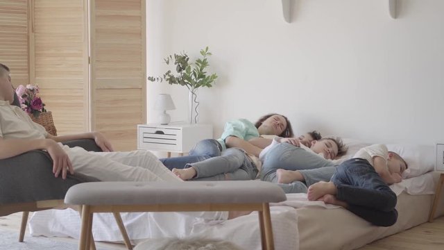 Young mother with her teen sons sleeping together in the big bed. One boy sitting on the chair. Happy loving family concept. Quiet hour in the family with many kids