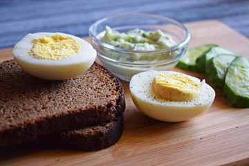 Boiled egg with black bread and fresh cucumbers on wooden background.