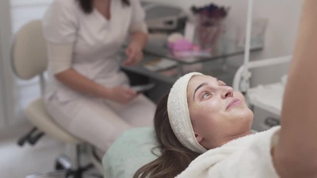 Pretty female client lying on the couch in beaty spa/salon. She is doing selfie while being covered with beauty mask. Girl's smiling a lot.