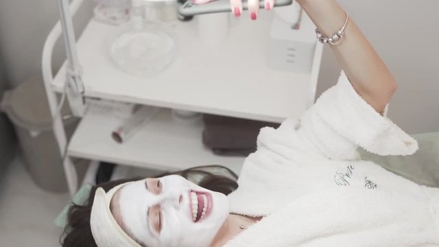 Pretty female client lying on the couch in beaty spa/salon covered with white mask. Girl making selfies, duck faces, sticking the tongue, winking, rolling eyes, showing teeth, sending air kiss