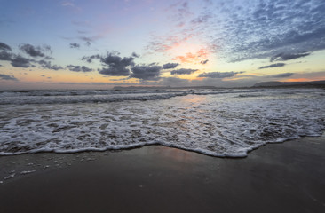 Location place Agia Marina Beach, island Crete, Greece. Sea coast spangled by rocks, the sunrise is reflecting on the wet sand. The mountains in haze on the horizon. Storm sea with high waves.