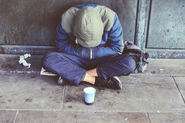 Homeless adult man sitting on the street in the shadow of the building and begging for help and...