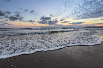 High waves with foam spread on the sand on the coast. The light of unbelievable sunset is reflecting on the sea. Pre dawn time. Beautiful enlighten sky with clouds. Mountains. Romantic relax place.