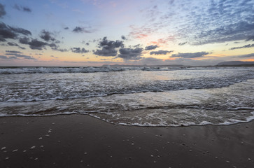 High waves with foam spread on the sand on the coast. The light of unbelievable sunset is reflecting on the sea. Pre dawn time. Beautiful enlighten sky with clouds. Mountains. Romantic relax place.