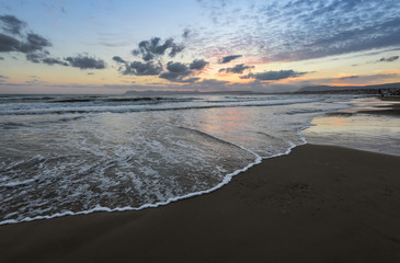 High waves with foam spread on the sand on the coast. The light of unbelievable sunset is reflecting on the sea. Pre dawn time. Beautiful enlighten sky with clouds. Mountains. Romantic relax place.