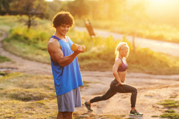 Smiling muscular man in sportswear doing exercises with dumbbells in nature on sunny summer day. In background woman warming up.