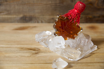 maple syrup in a glass bottle in the form of a maple leaf on ice from large ice cubes on a wooden rustic background with copy space for text