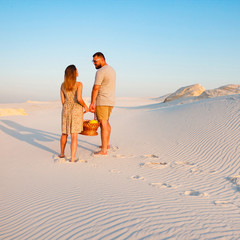 lovely attractive couple on the white sand beach or in the desert or in the sand dunes, guy and a girl with a basket in their hands, the couple left traces or footprints on the sand, rear view