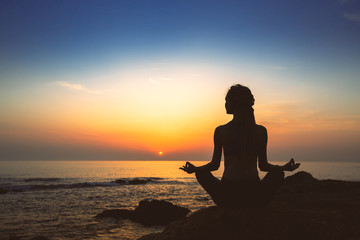 Silhouette of woman yoga in Lotus position on the shore of ocean at evening.