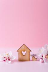 Closeup wooden house with hole in form of heart surrounded by white flowering tree branches on pastel pink background.