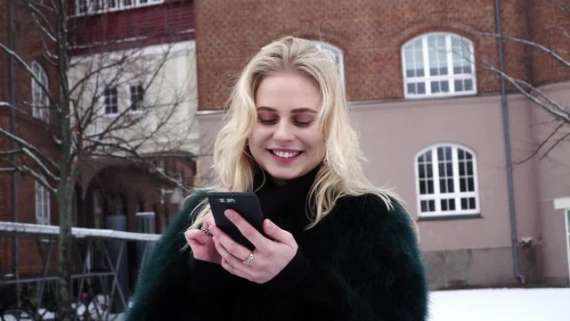 Happy and beautiful young woman is swiping on the screen of her smartphone outdoors in a winter time.
