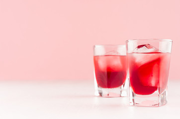 Fresh red strawberry cocktails with ice cubes in two elegant misted shot glasses on soft light pink color background and white wooden table, copy space