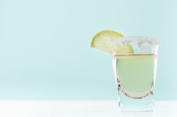 Elegant golden tequila in shot glass with rim of salt and slice lime on pastel mint color background, copy space.