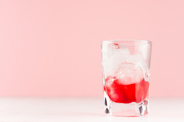 Strawberry red cocktail in shot glass with ice cubes on white wood table and pastel pink wall, copy space.
