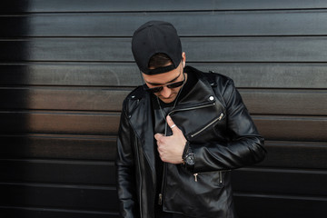 Fashionable young hipster man in sunglasses in a vintage leather jacket in a stylish baseball cap...