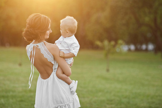 Beautiful Brunette Model Girl, Young Mom In Short White Stylish Dress Holding On Her Hands Her Cute Happy Baby Boy And Posing At The Green Park At The Sunset