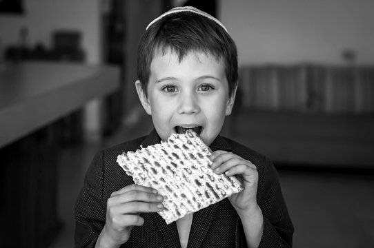 Cute Caucasian child in a yarmulke taking a bite from a traditional Jewish matzo unleavened bread in a room. Black and white image.