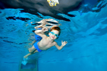 Sporty little boy swims underwater in the pool near the surface of the water, looking and posing for the camera. Portrait. Underwater photography. Horizontal orientation